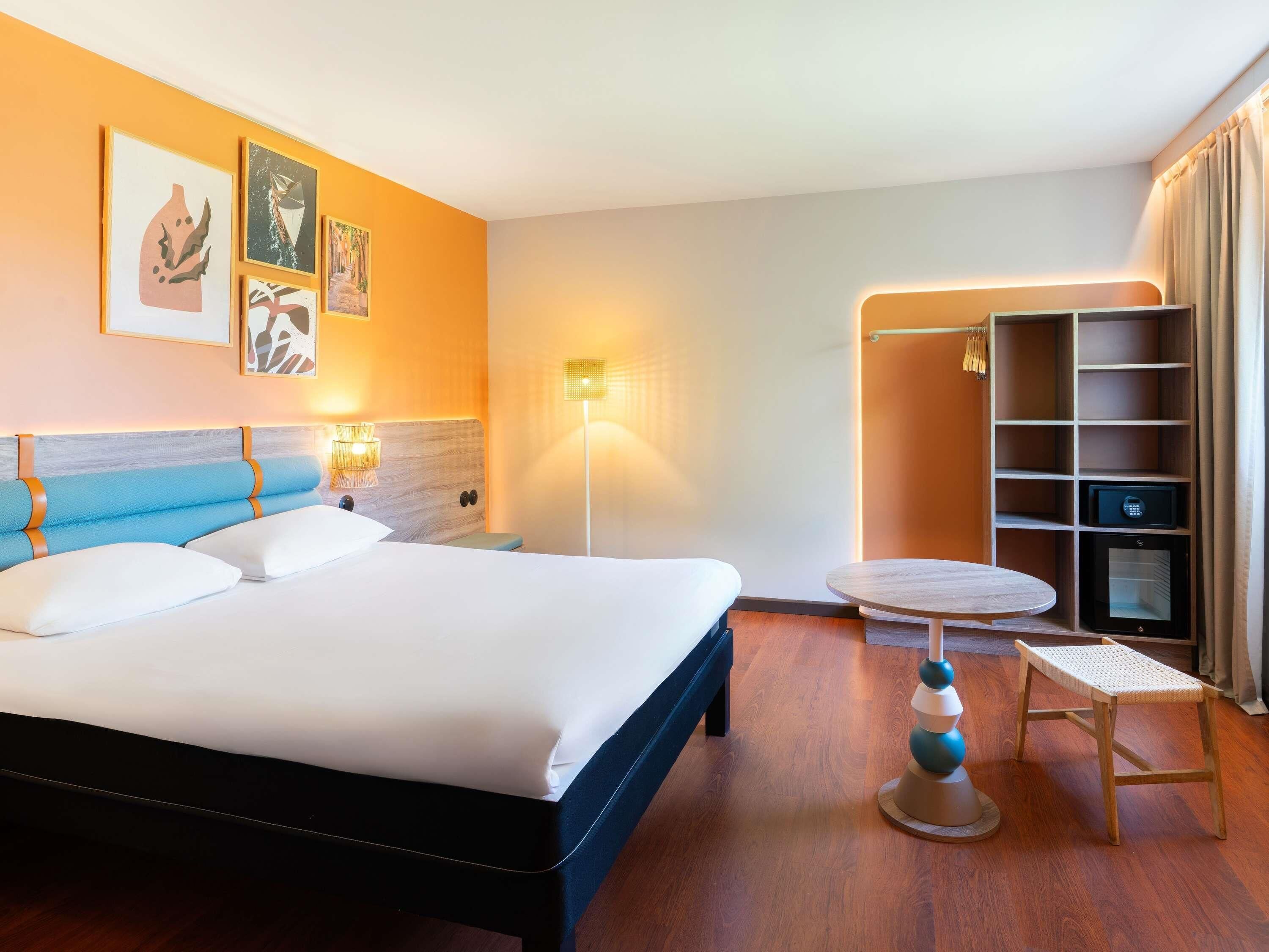 A hotel room with a bed, bedside lamps, a round table, a chair, a wardrobe, and a wall with artwork.