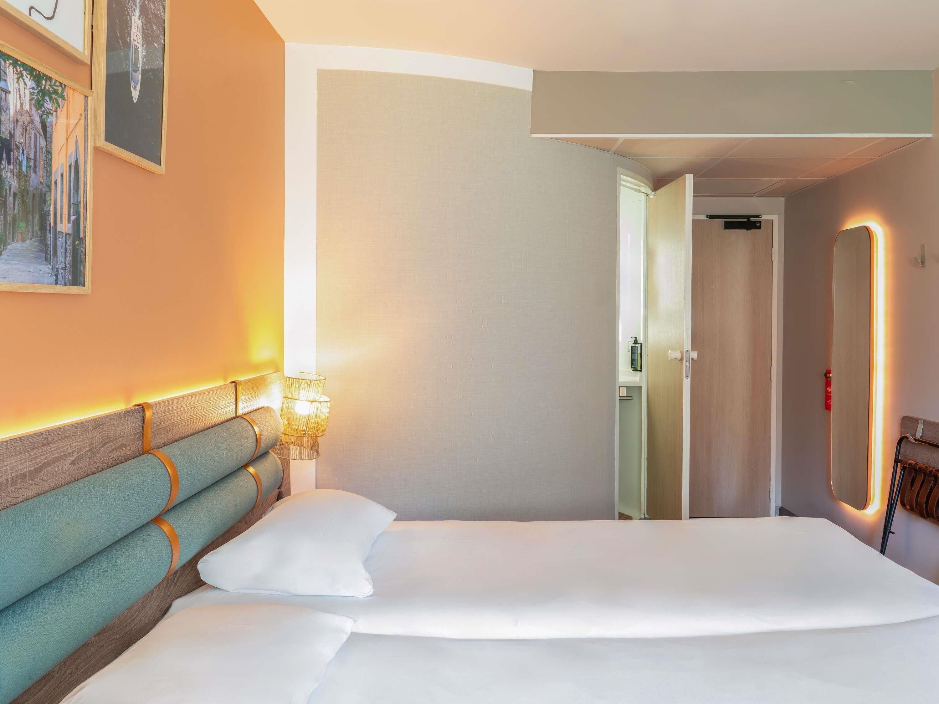 Une chambre d’hôtel avec un grand lit, une tête de lit en bois, une lampe fixée au mur et une porte donnant sur une autre pièce.