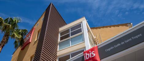 A hotel with a modern design, featuring a prominent red 'ibis' sign and a 'Cogolin Golf de Saint-Tropez' sign.