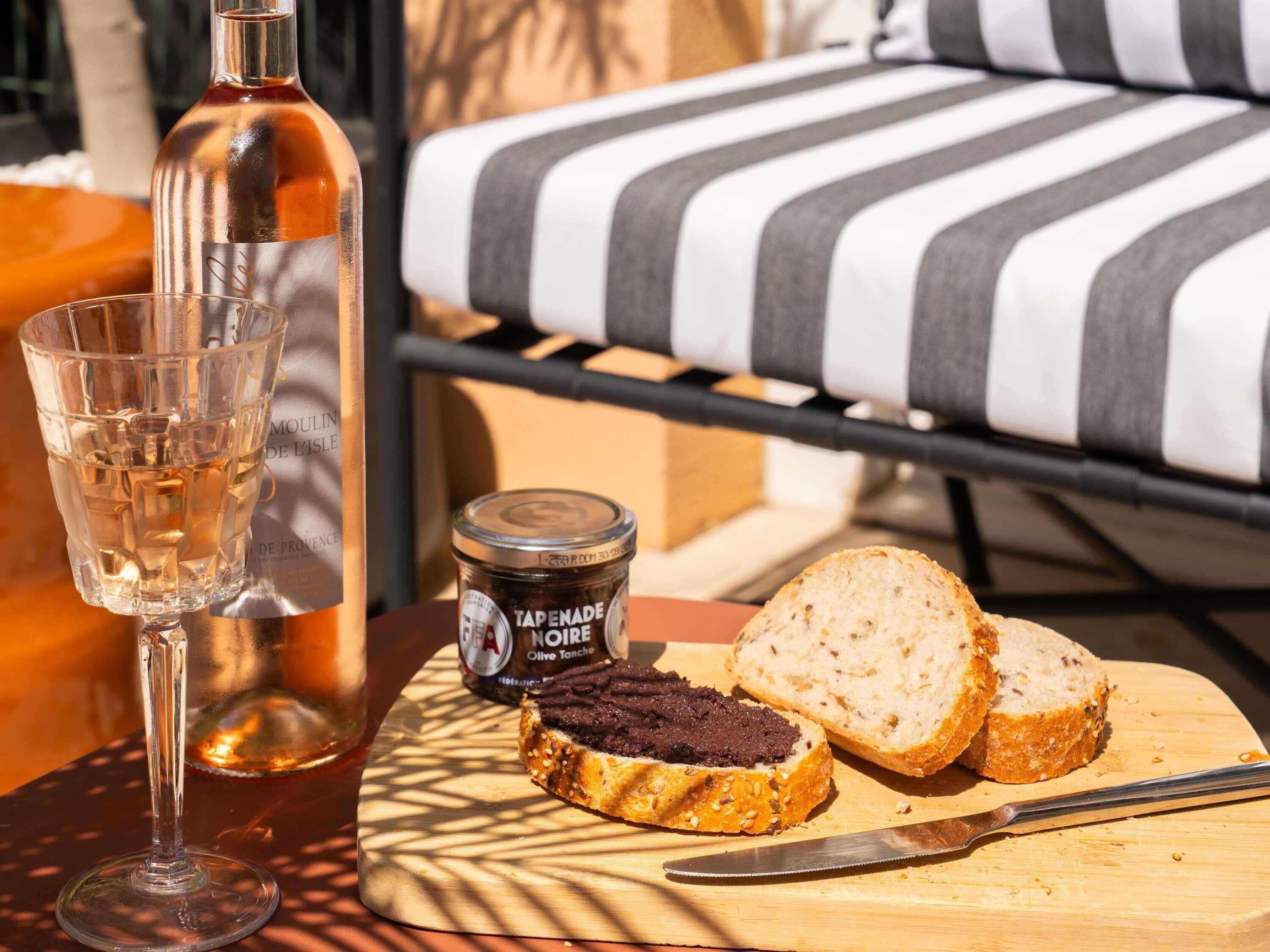 Una mesa con una botella, una copa, un tarro de tapenade y rebanadas de pan sobre una tabla de madera.