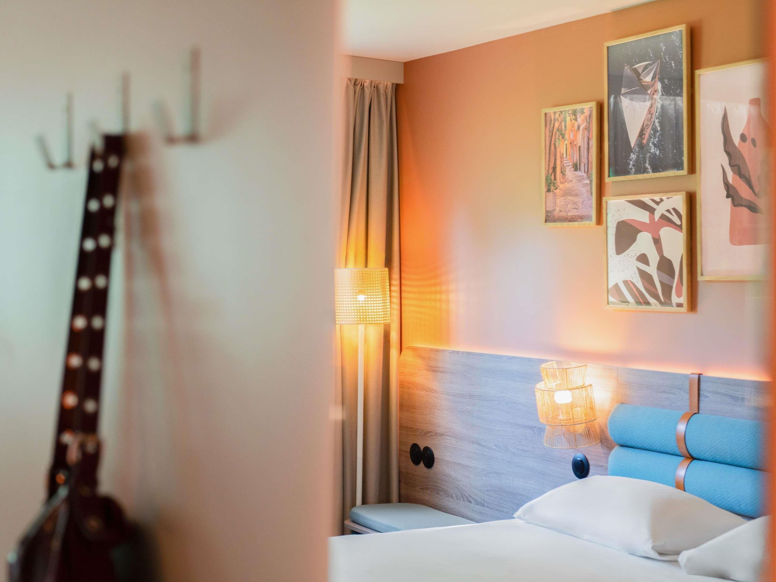 Une chambre d’hôtel avec un lit, une tête de lit, une lampe fixée au mur et une guitare appuyée contre le mur.