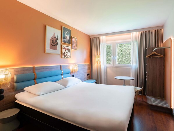 Habitación de hotel con una cama grande, lámparas de noche, una mesita, perchero y ventana con cortinas.