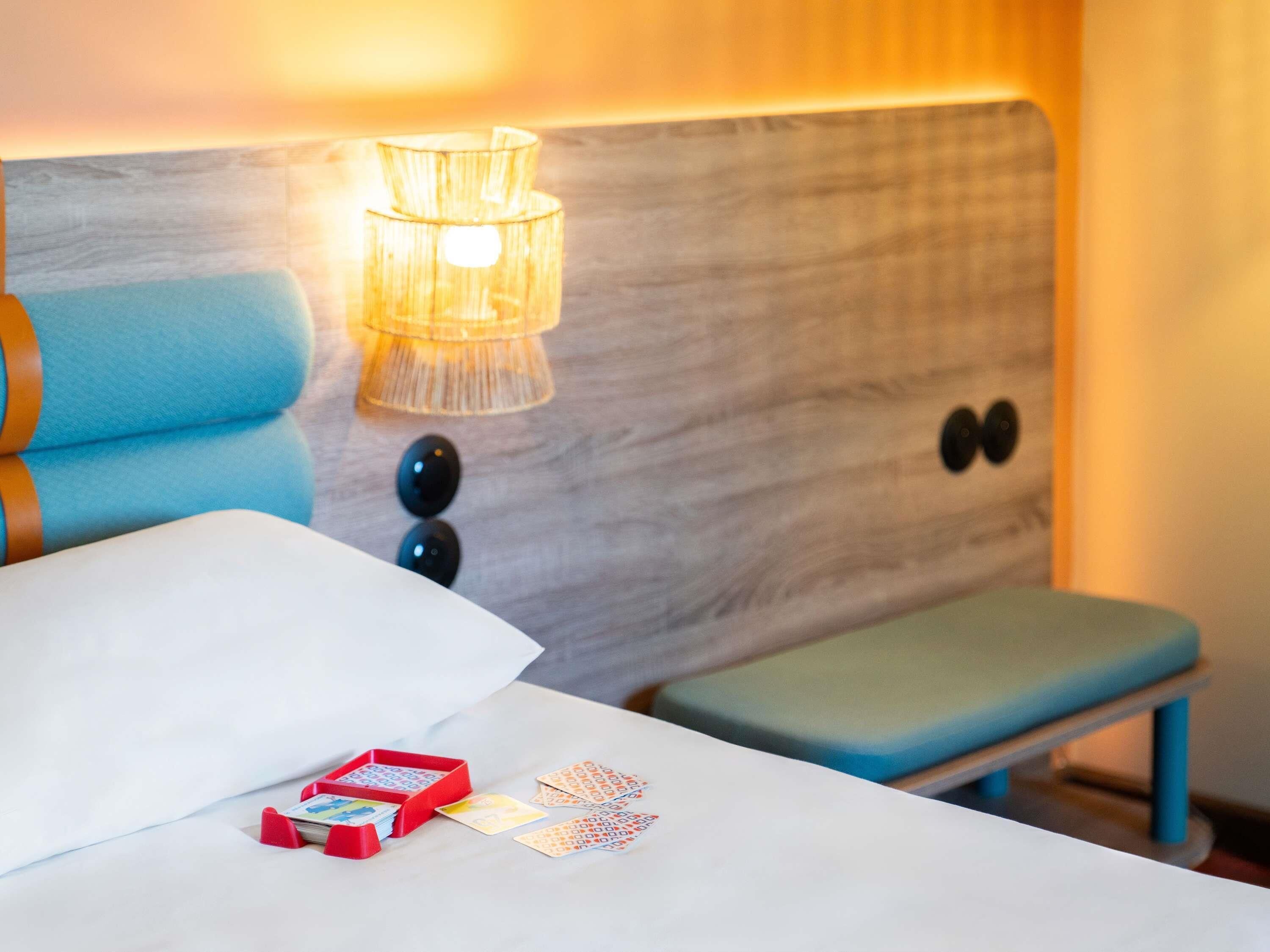 A hotel room with a bed, a bedside table with a lamp, a bench, and a small table with a box and cards.
