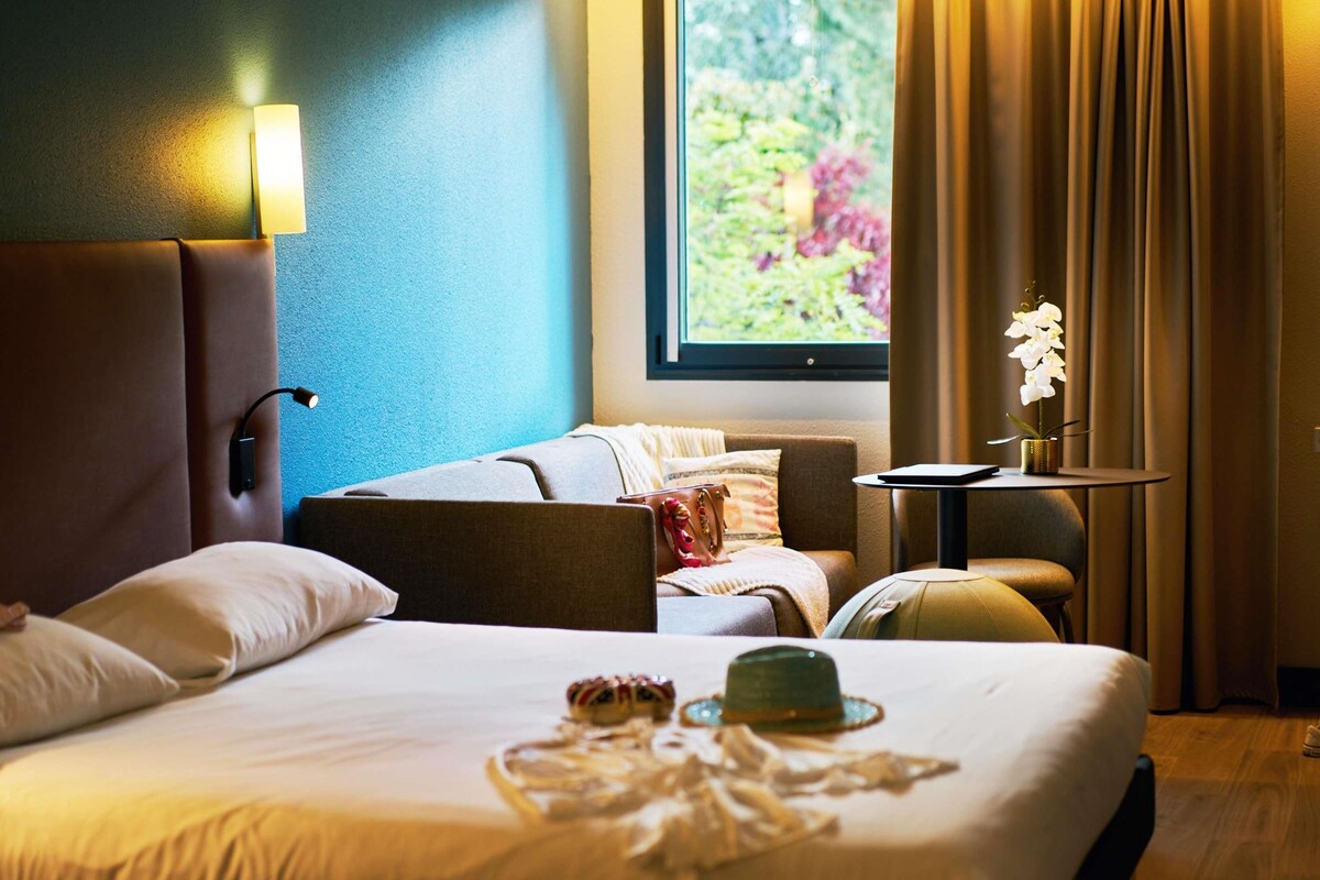 A hotel room with a large bed, a bedside table, a lamp, a window with curtains, and a view of greenery outside.