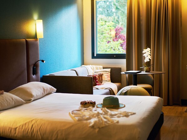 A hotel room with a large bed, a bedside table, a lamp, a window with curtains, and a view of greenery outside.