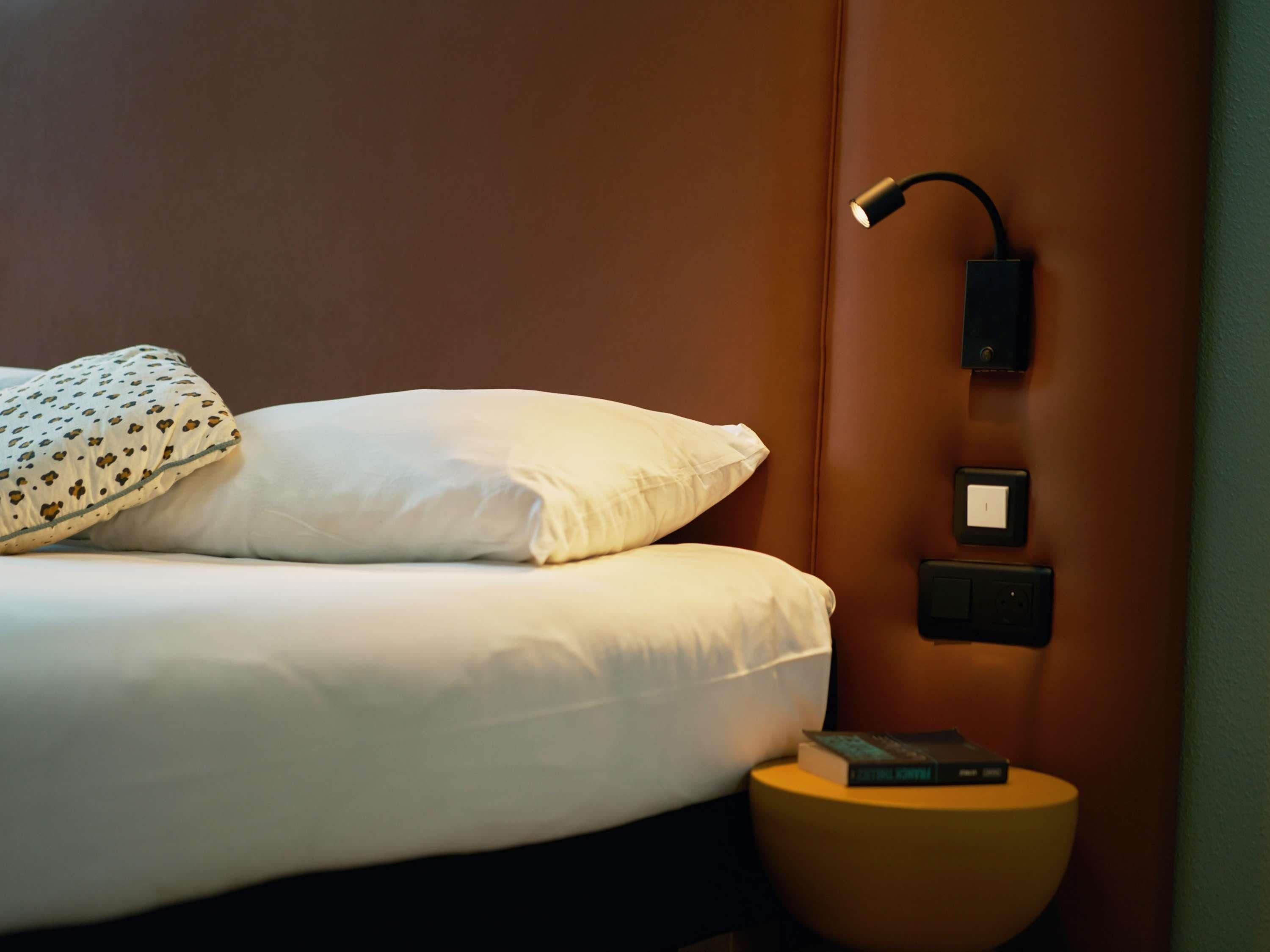 Un lit bien fait, avec du linge de lit blanc et un coussin à motifs, une lampe de chevet, une petite table de chevet avec un livre, et un interrupteur fixé au mur.