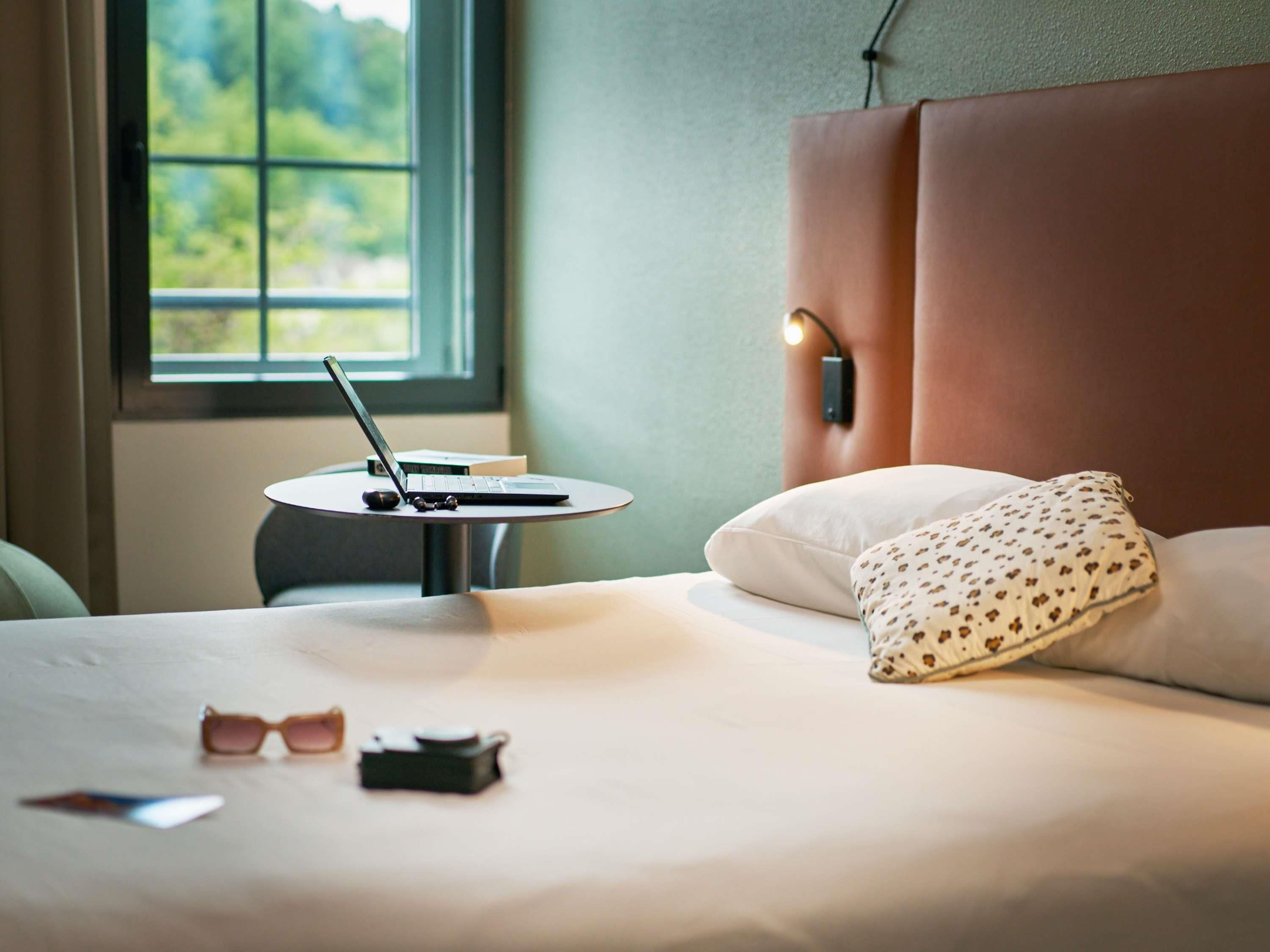 Een net opgemaakt bed met een kussen, een laptop op een klein tafeltje ernaast, en een paar zonnebrillen op het bed.