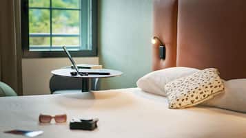 Una cama bien hecha con una almohada, un ordenador portátil sobre una mesita auxiliar cercana y un par de gafas de sol sobre la cama.