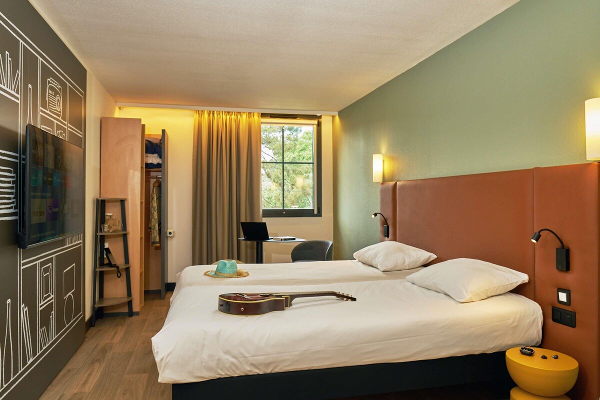 A hotel room with a bed, a guitar, a laptop, and a desk with a chair.