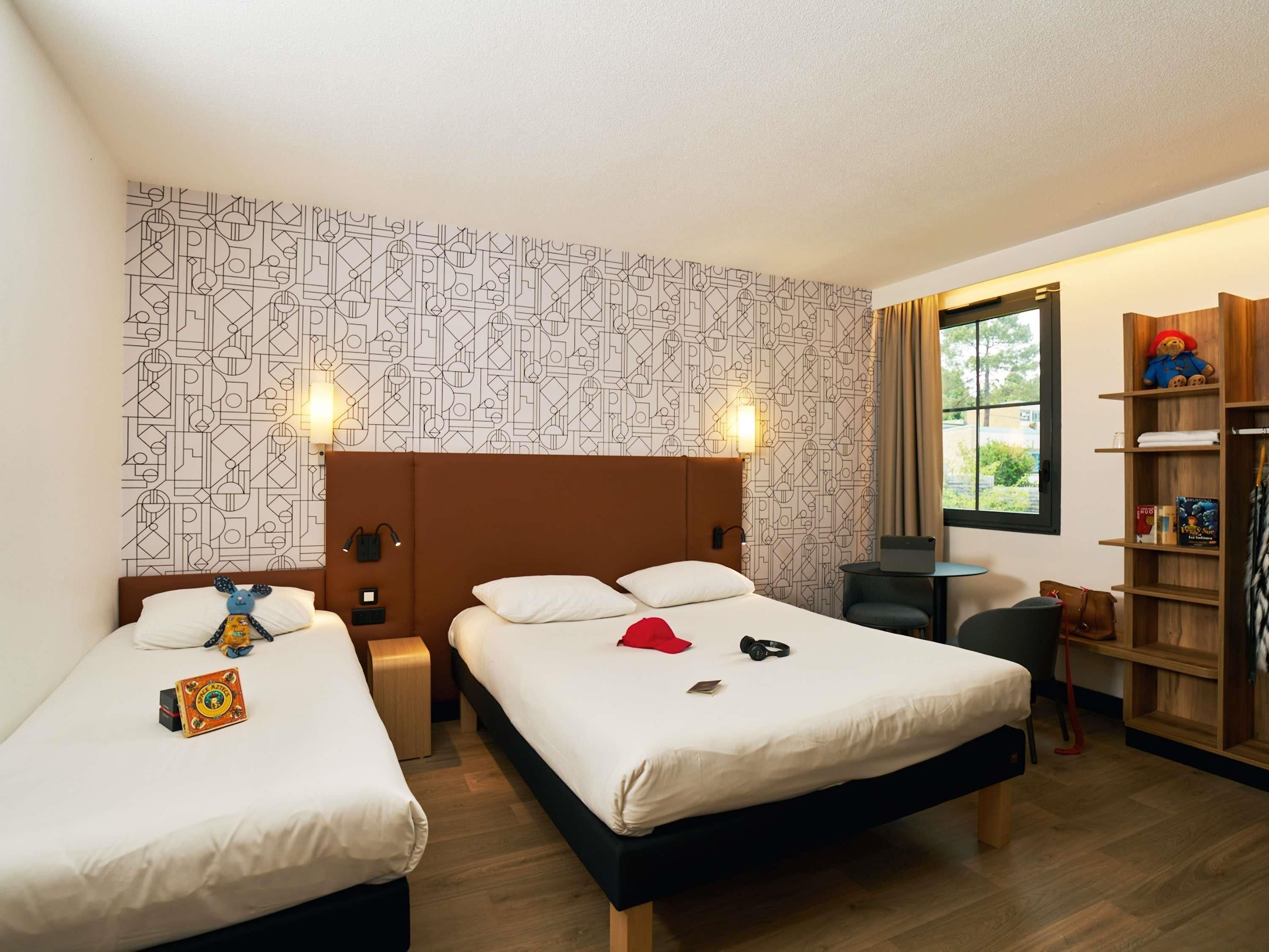 A hotel room with two beds, a desk, a chair, a bookshelf, and a window with a view.