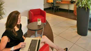 A woman using a laptop in a modern lobby with red seating, a map on the wall, and a large potted plant. - ibis Sarlat (Sarlat-la-Canéda)
