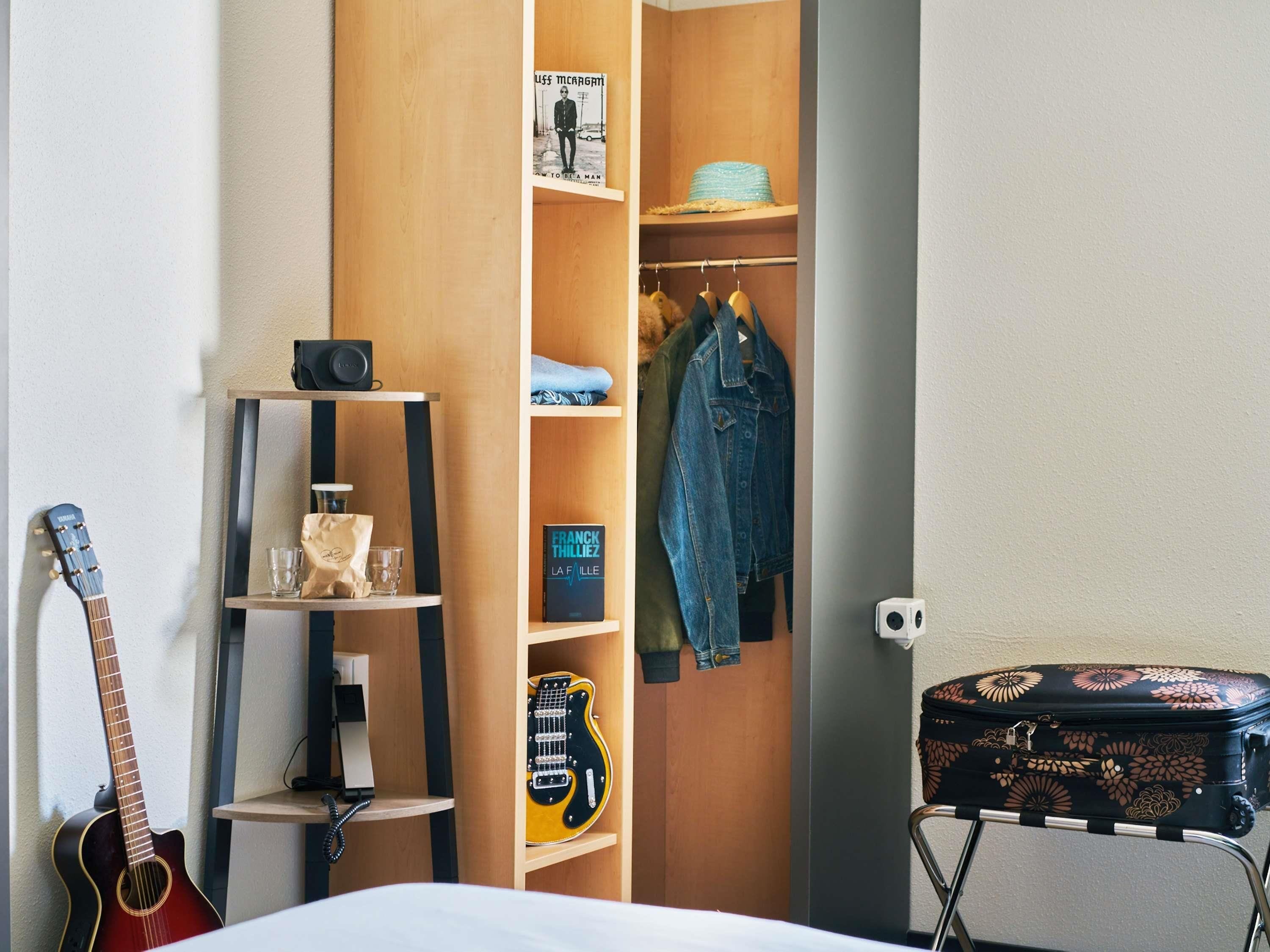 A room with a guitar, a suitcase, a shelf with a book, and a closet with hanging clothes.