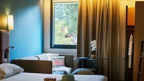 Habitación de hotel con cama, escritorio, silla, lámpara, ventana con cortinas y vistas al exterior.