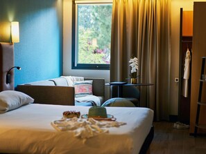 A hotel room with a bed, a desk, a chair, a lamp, a window with curtains, and a view of greenery outside. - ibis Sarlat (Sarlat-la-Canéda)