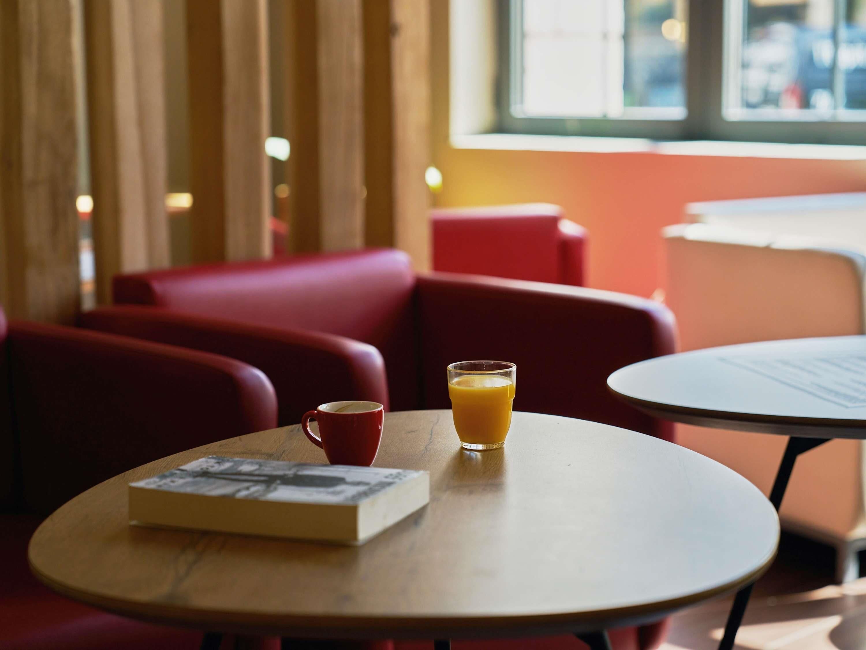Una mesa con un libro, un vaso de jugo de naranja y una taza roja, con una ventana al fondo.