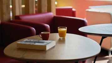 Una mesa con un libro, un vaso de jugo de naranja y una taza roja, con una ventana al fondo.