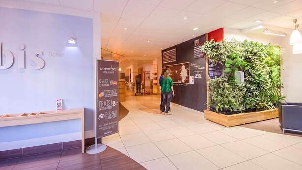 A modern interior with a 'bis' sign, a wooden bench, a green wall with plants, and a tiled floor. - ibis Sarlat (Sarlat-la-Canéda)