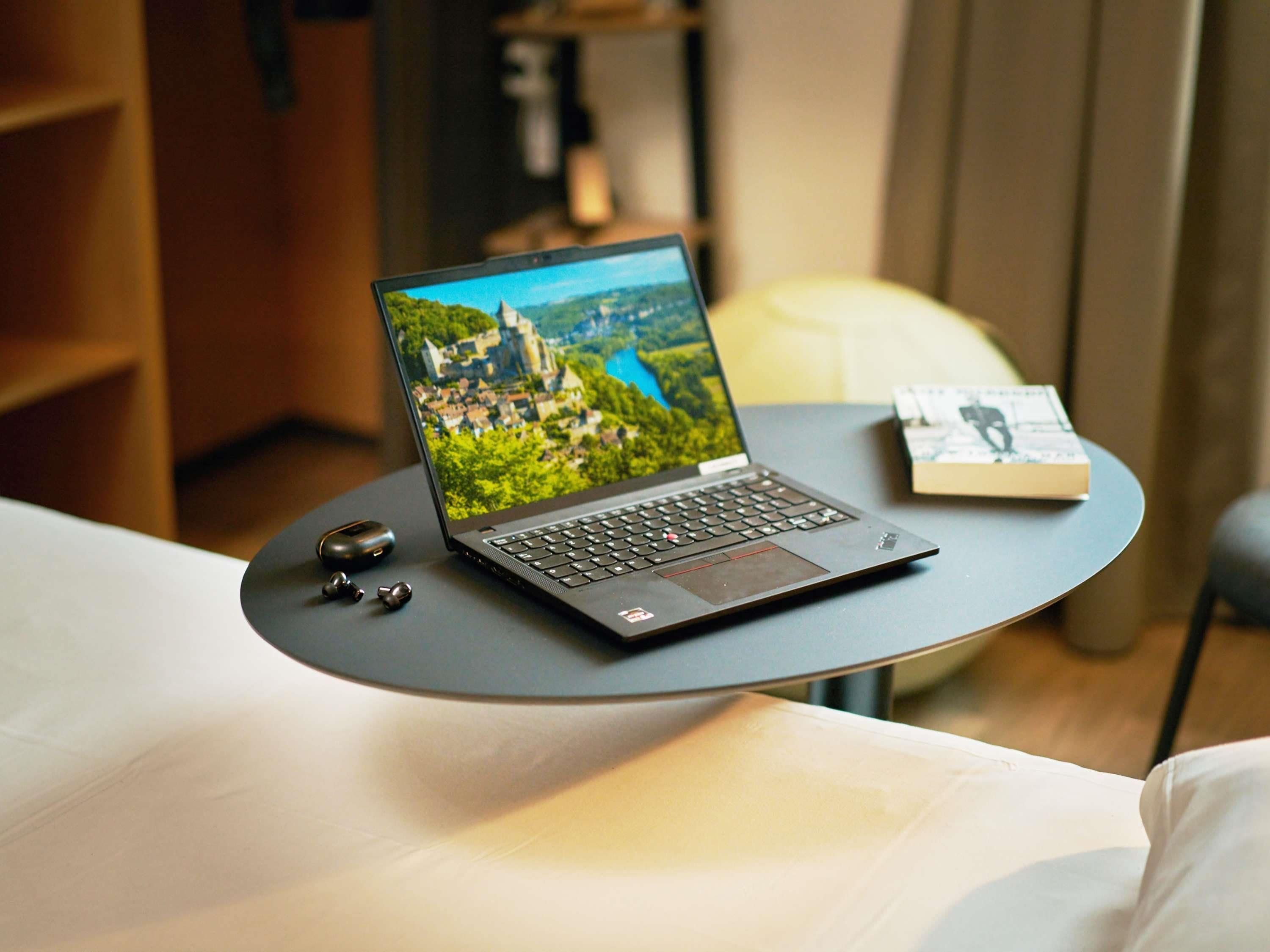 A laptop on a small table with a book beside it, in a room with a bed and a chair.