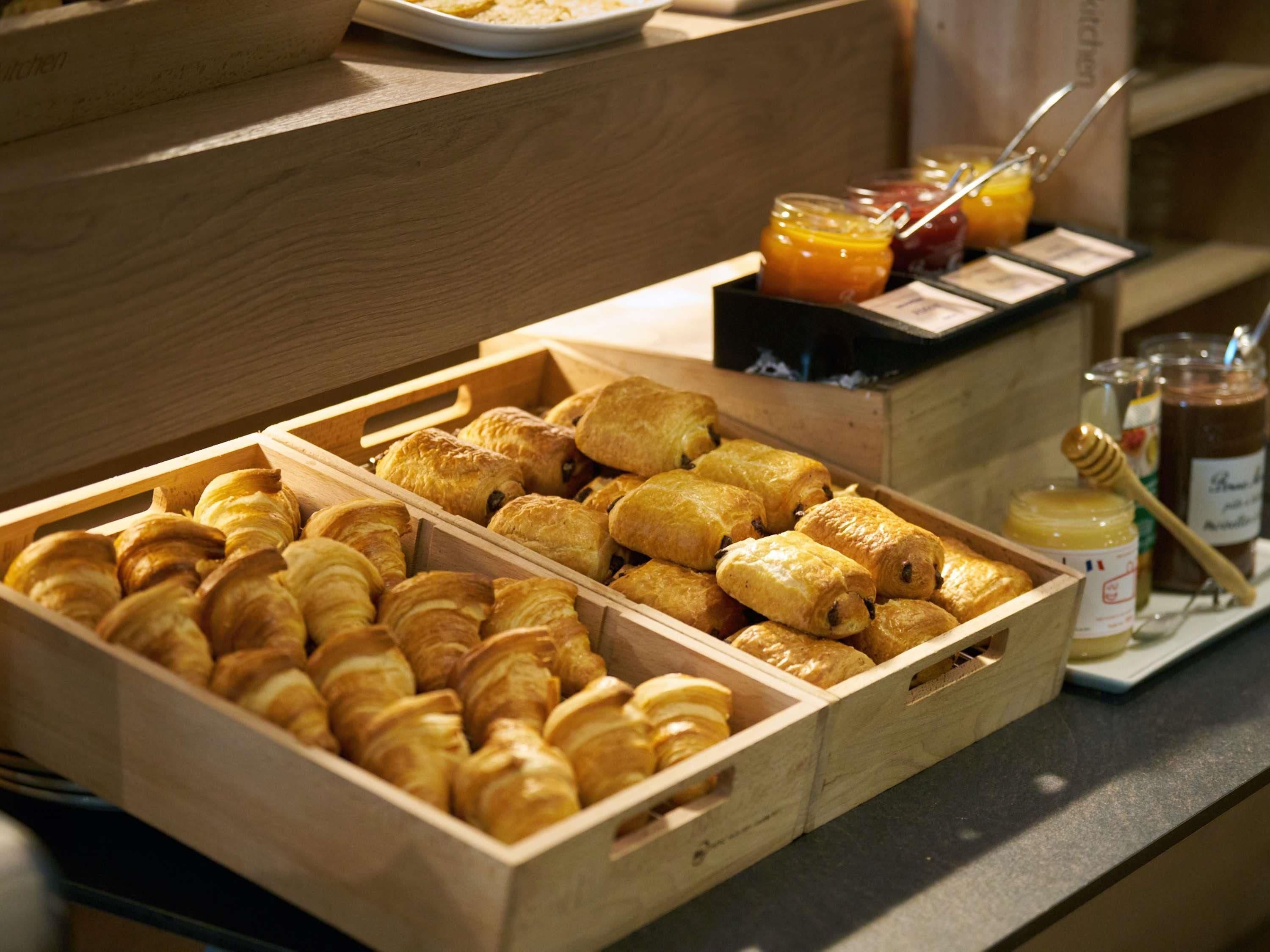 Paneras de madera llenas de croissants y pasteles, junto con una variedad de mermeladas y miel sobre una encimera.