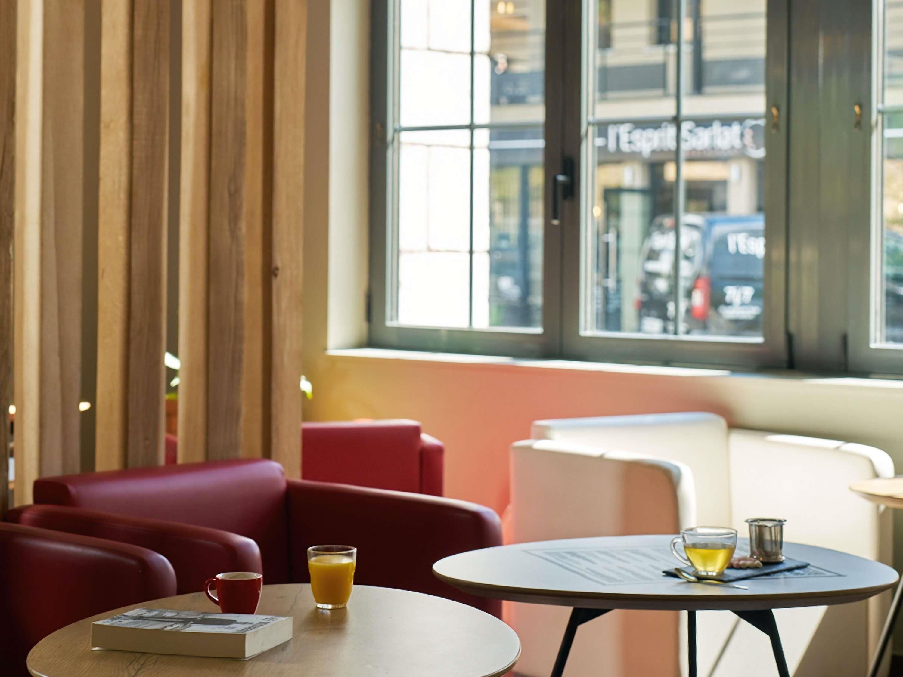 Un coin salon avec une table, un verre de jus d’orange et une tasse, donnant sur une rue et un immeuble avec une enseigne à travers une fenêtre.