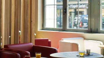Un coin salon avec une table, un verre de jus d’orange et une tasse, donnant sur une rue et un immeuble avec une enseigne à travers une fenêtre.