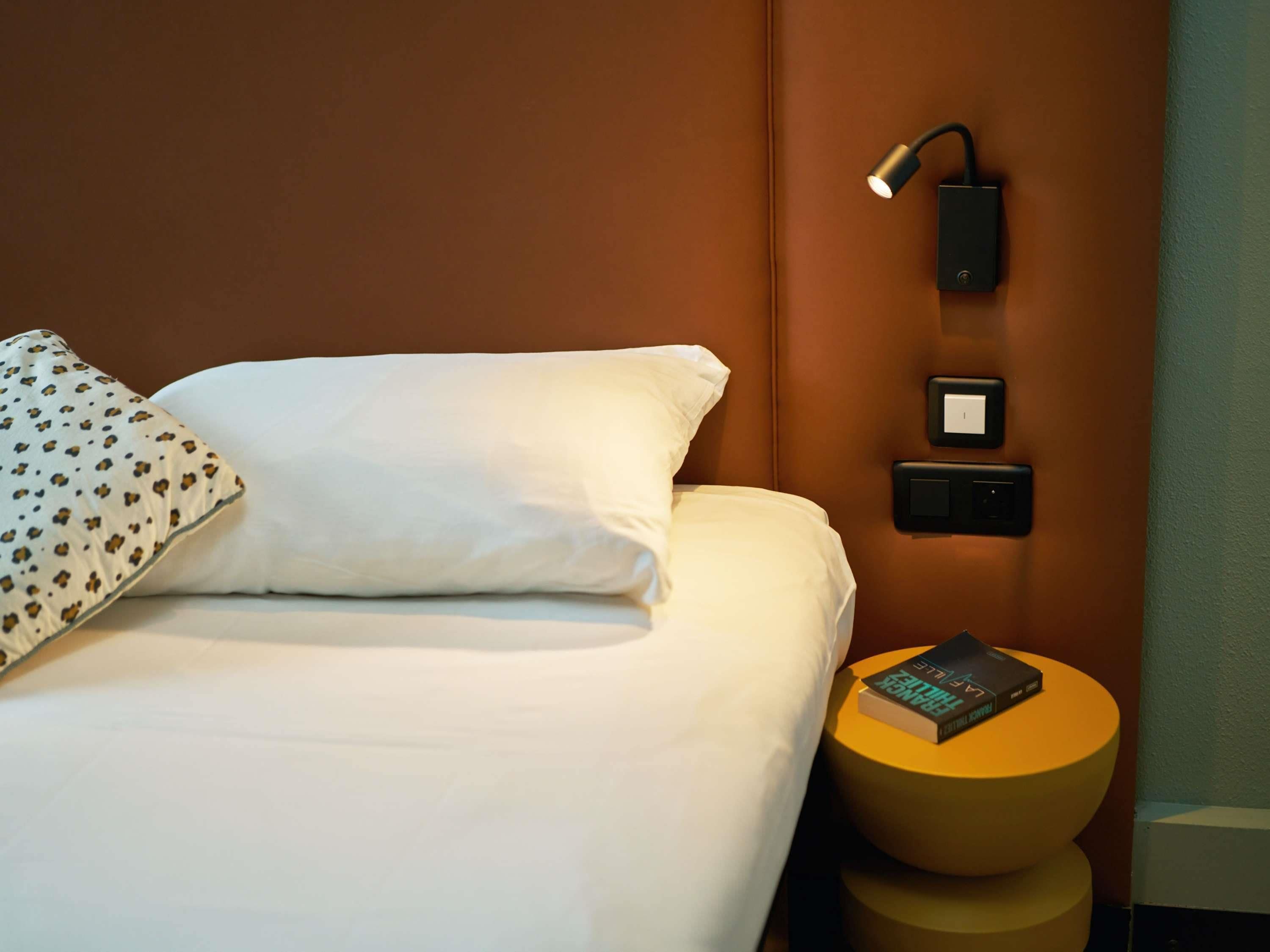 Hotelkamer met een bed, een bedlamp, een stopcontact en een klein, rond tafeltje met een boek.