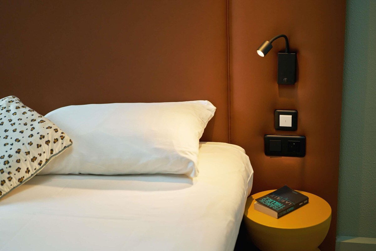 A hotel room with a bed, a bedside lamp, a power outlet, and a small round table with a book.