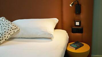 Hotelkamer met een bed, een bedlamp, een stopcontact en een klein, rond tafeltje met een boek.