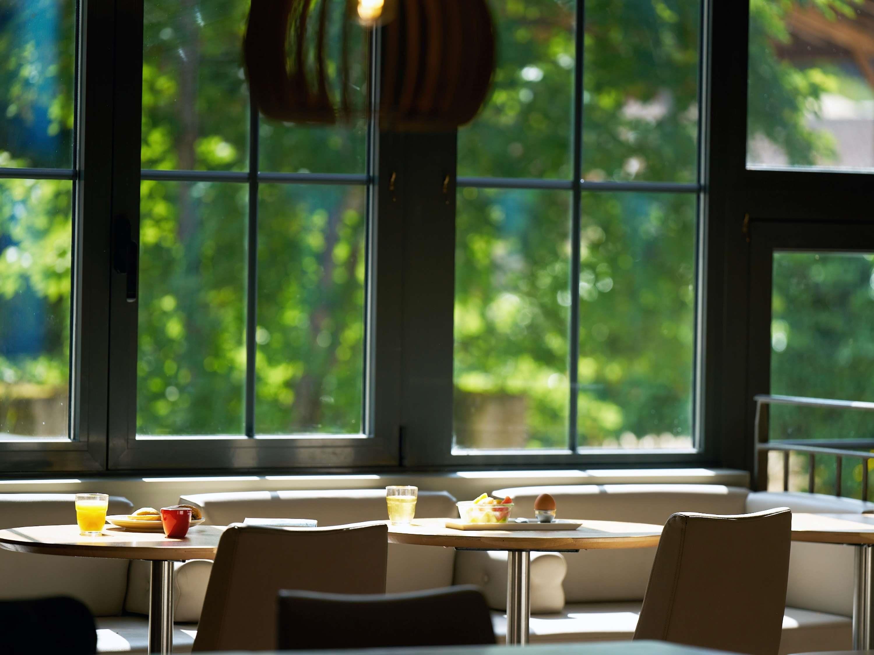 Zona pranzo con tavoli e sedie, ampie finestre che si affacciano sul verde e una lampada a sospensione.