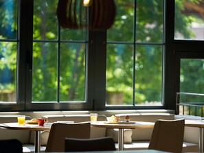 A dining area with tables and chairs, large windows with a view of greenery, and a hanging light fixture. - ibis Sarlat (Sarlat-la-Canéda)