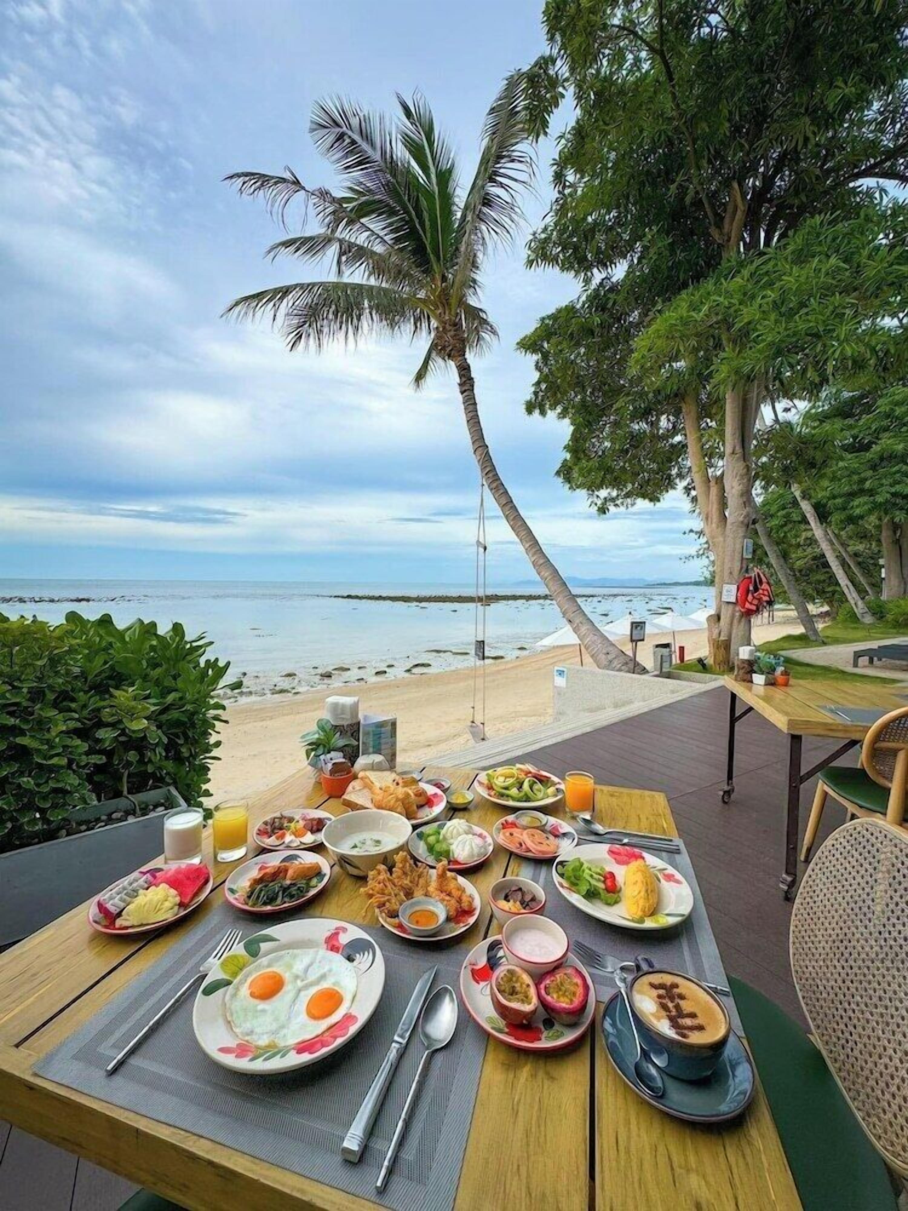 Mesa preparada para el desayuno, con huevos, fruta y bebidas, con vista a una playa con palmeras y cielo despejado.