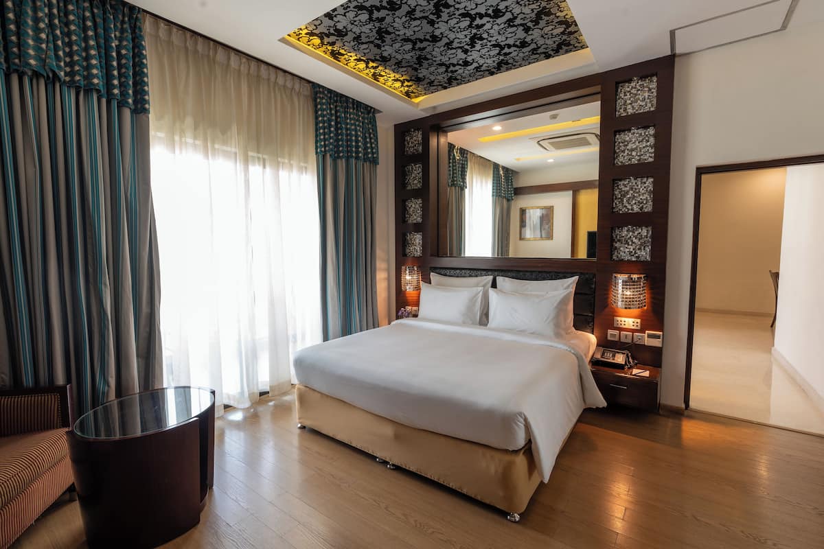 A modern hotel room with a large bed, a glass table, and a view of the city through the window.