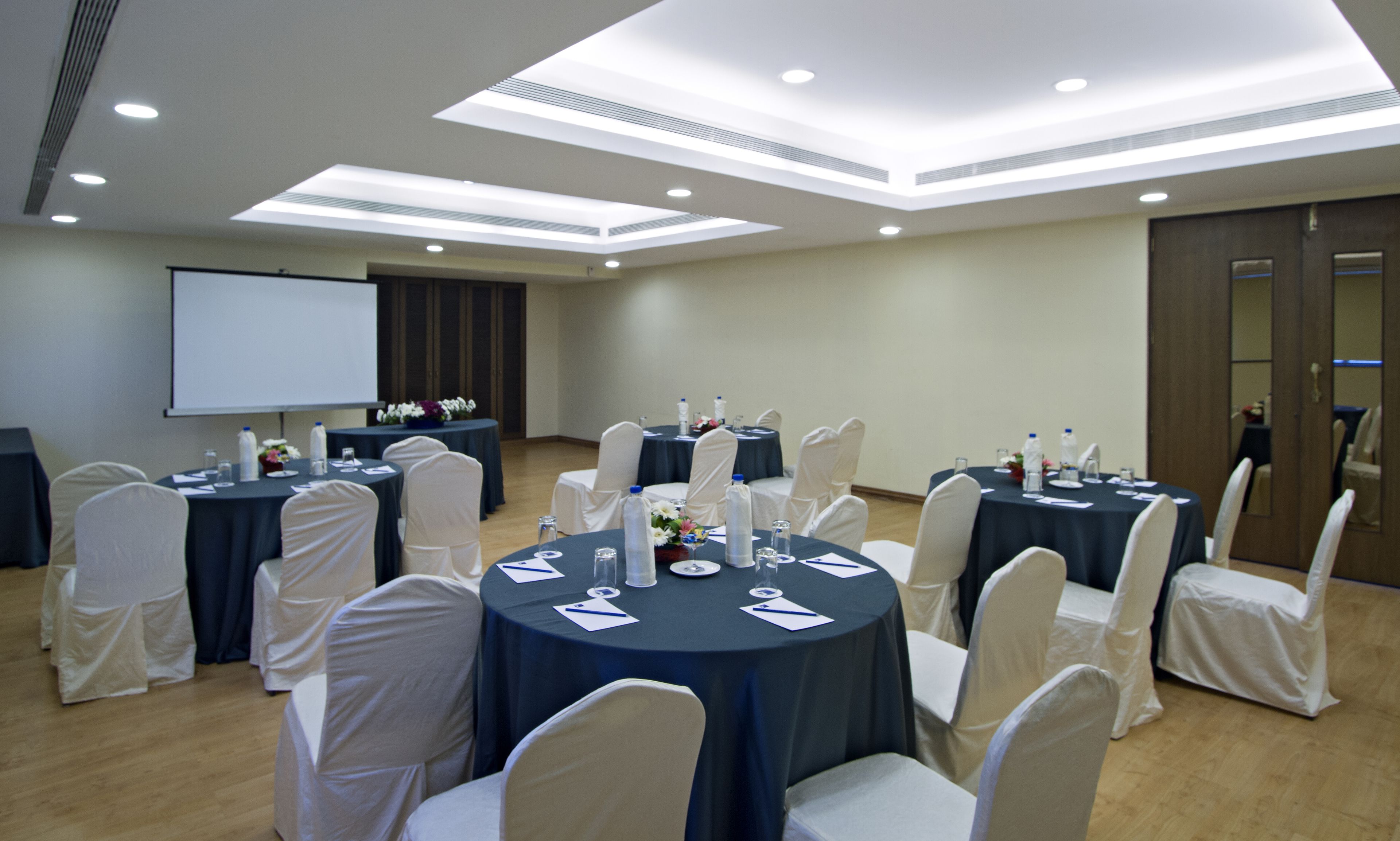 A conference room with round tables, white chairs, and a projector screen.