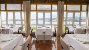 Un restaurante con mesas dispuestas para comer, amplias ventanas con vista al agua y al paisaje, y techo de madera.