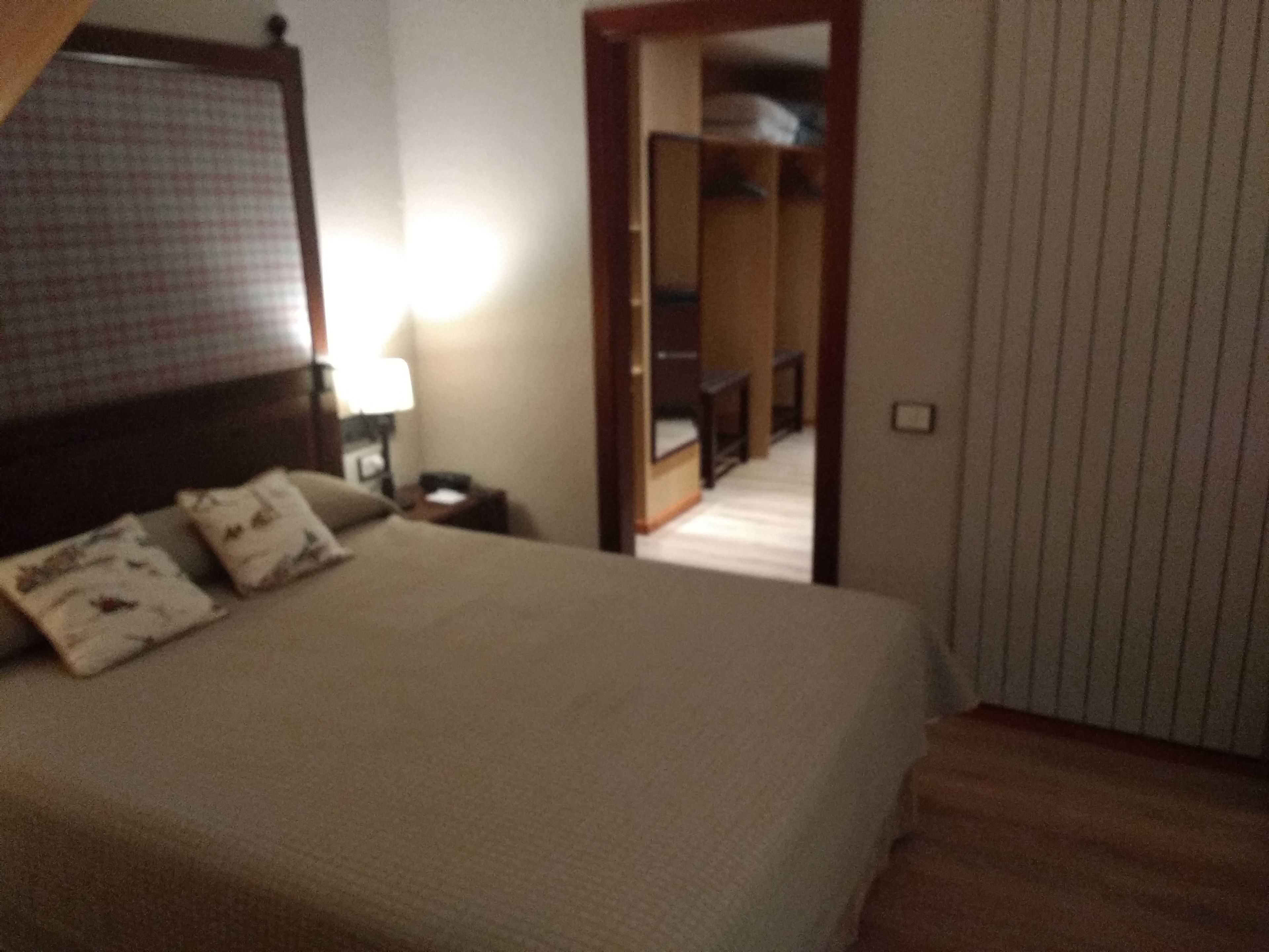 Un dormitorio con cama, almohadas, mesita de noche, lámpara y armario.