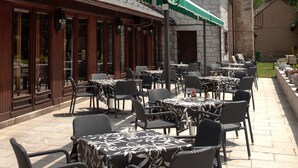 Un café al aire libre con mesas y sillas, un edificio de piedra con una torre, y un toldo verde.