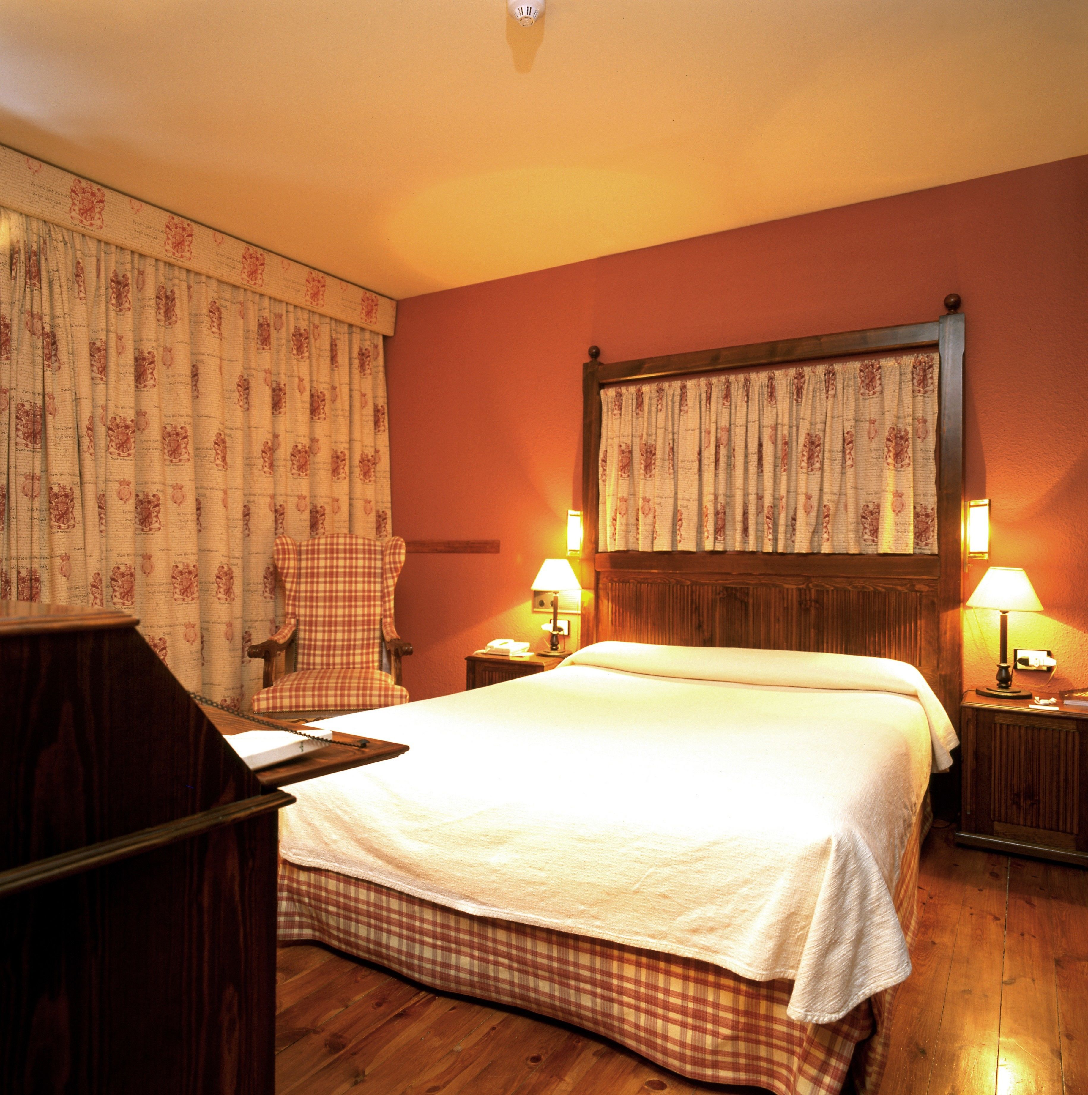 Une chambre d’hôtel avec un grand lit, deux lampes de chevet, une tête de lit en bois, une chaise à carreaux et un bureau en bois avec un téléphone.
