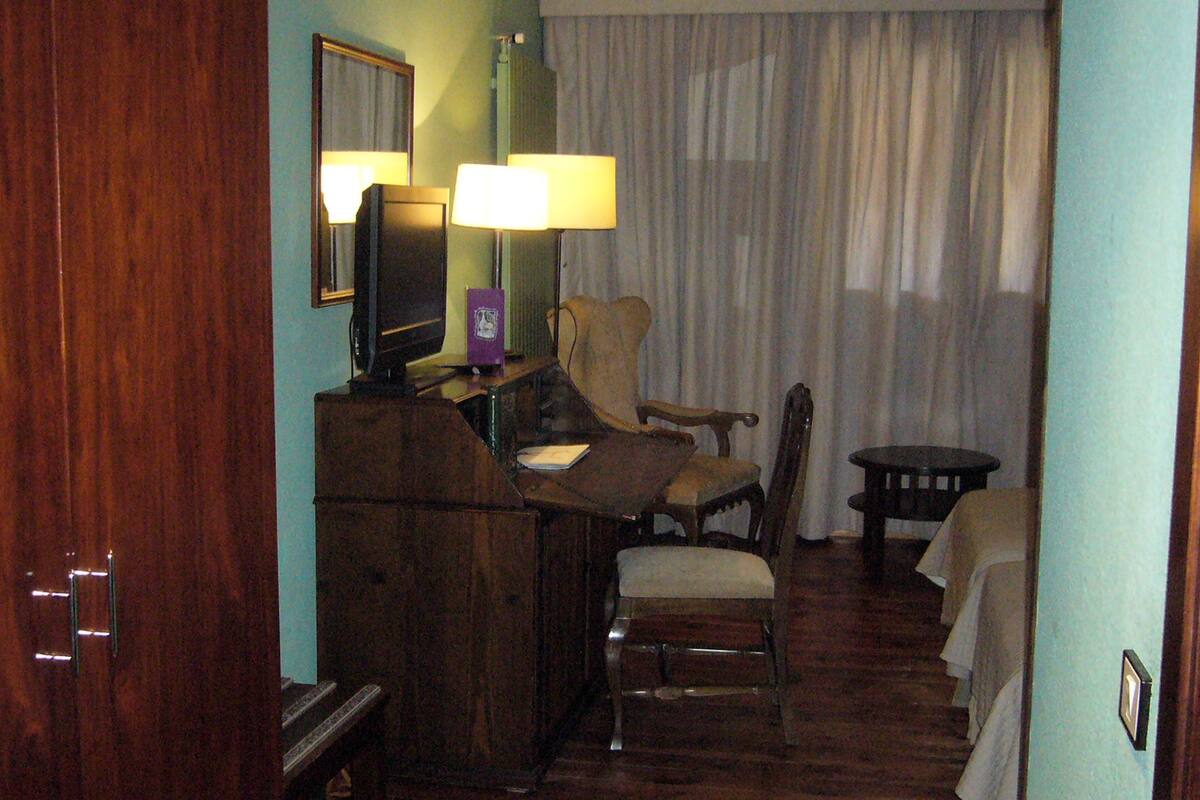 A hotel room with a wooden desk, a chair, a small table, a lamp, and a bed with a white comforter.