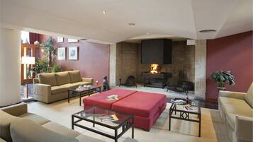 Una sala de estar con un sofá color beige, un reposapiés rojo y mesas de centro de vidrio. Hay una chimenea encendida, una pared de piedra y una planta.