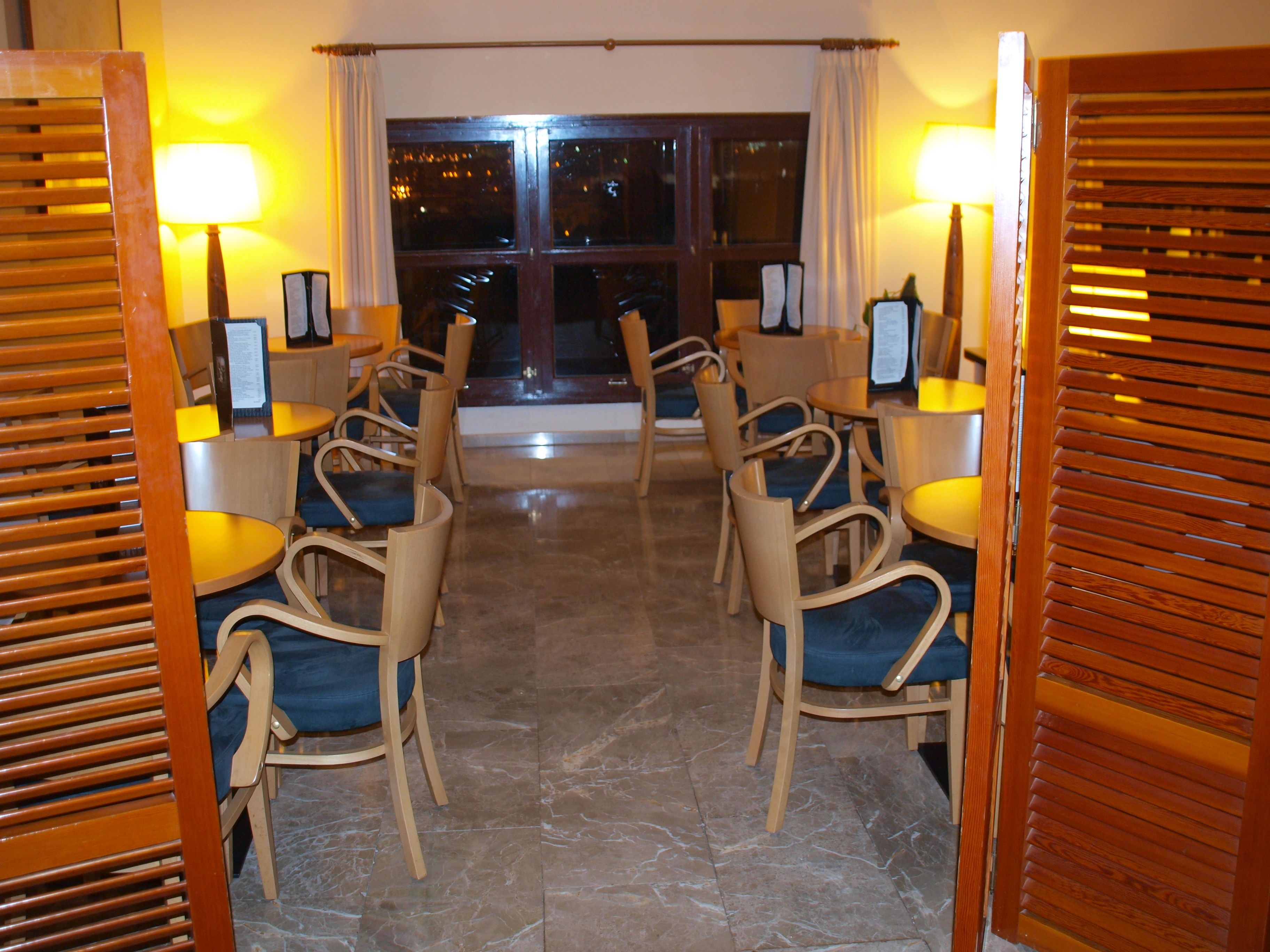 A dining area with wooden chairs and tables, illuminated by lamps, and a view of the night outside.
