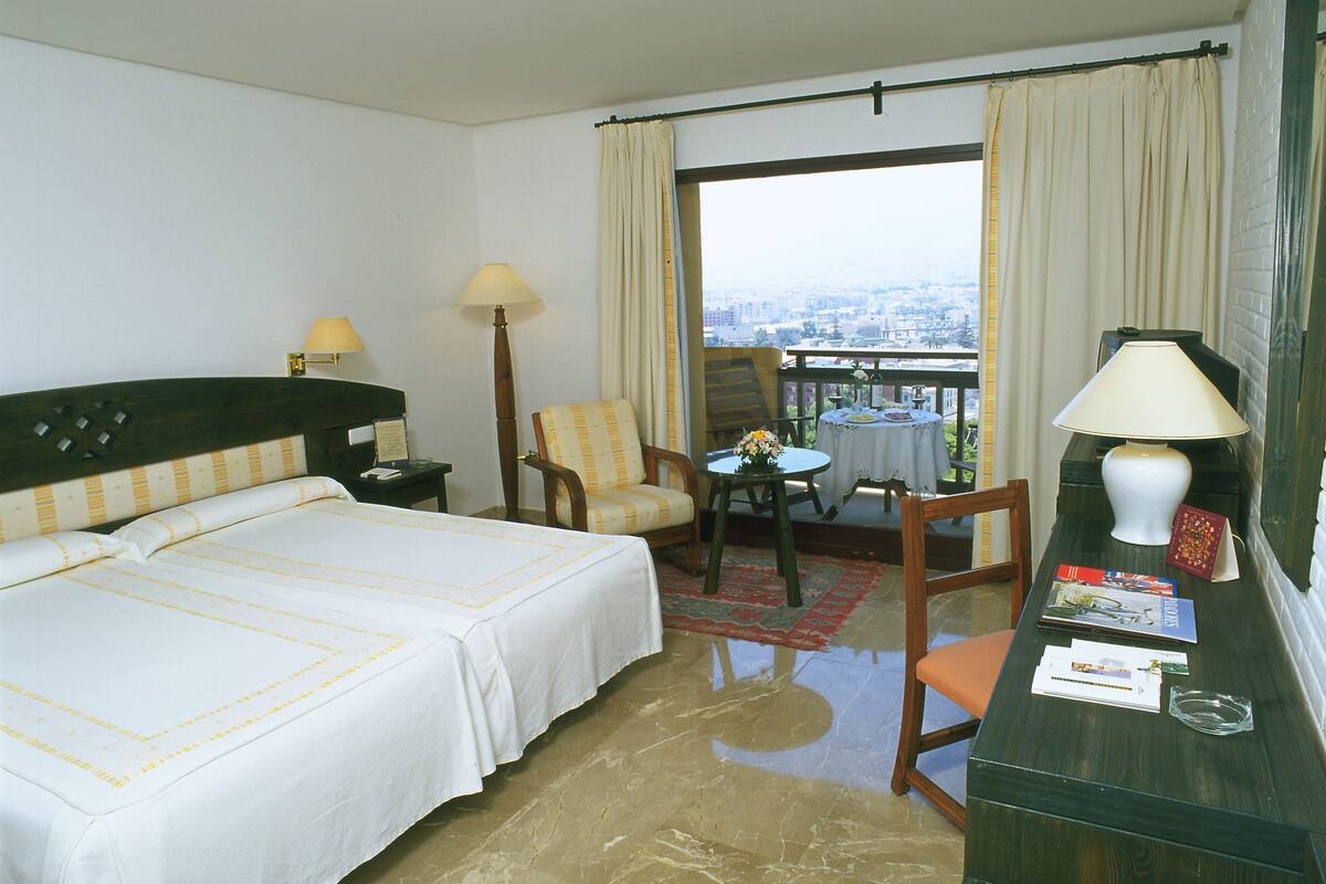 Une chambre d’hôtel avec deux lits, un bureau avec une lampe, une chaise, une petite table avec un vase et une vue sur la ville grâce à de grandes fenêtres.