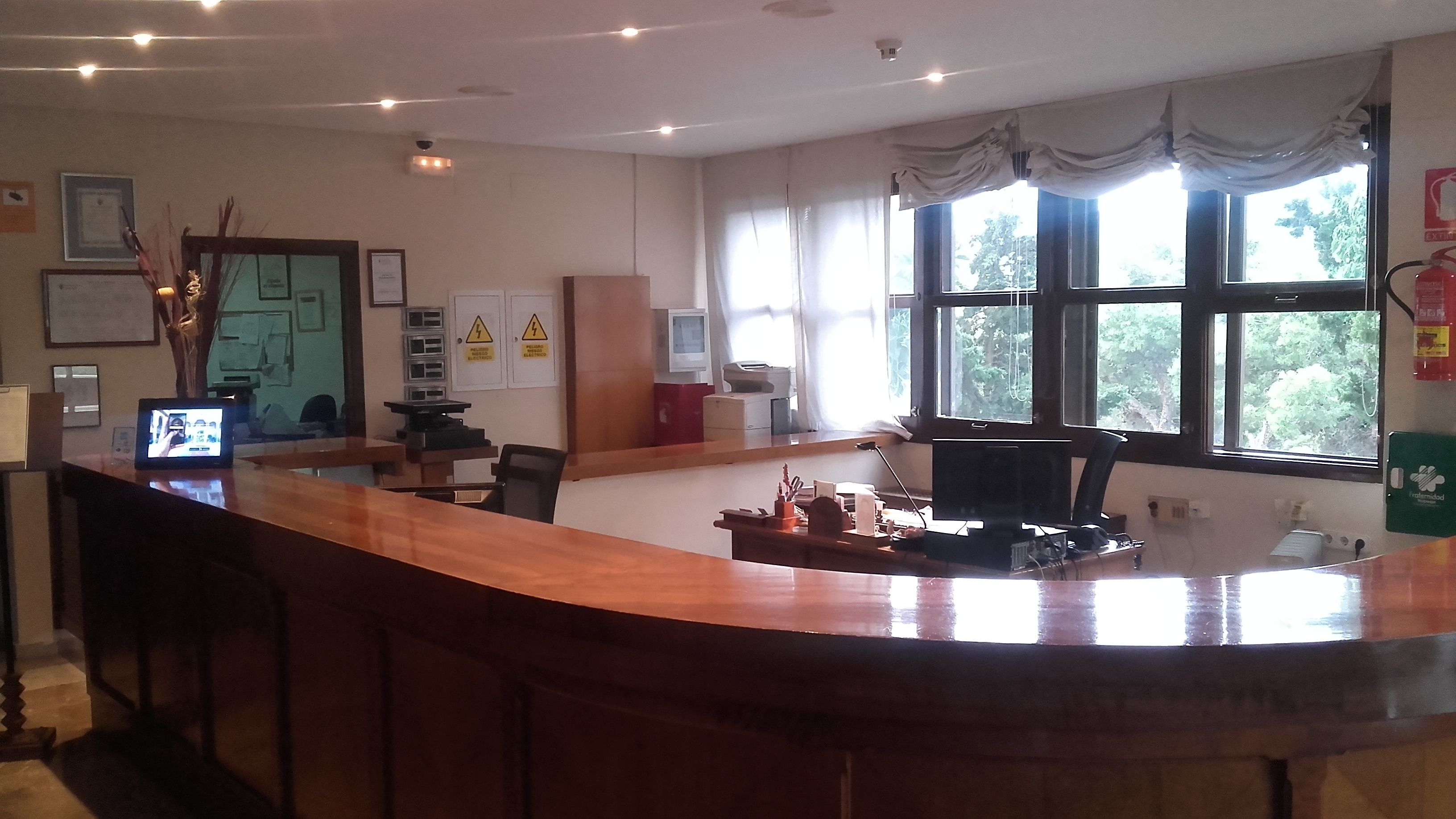 Una zona de recepción con un mostrador de madera, un monitor de computadora y un teléfono. Hay certificados enmarcados en la pared y una ventana con cortinas.