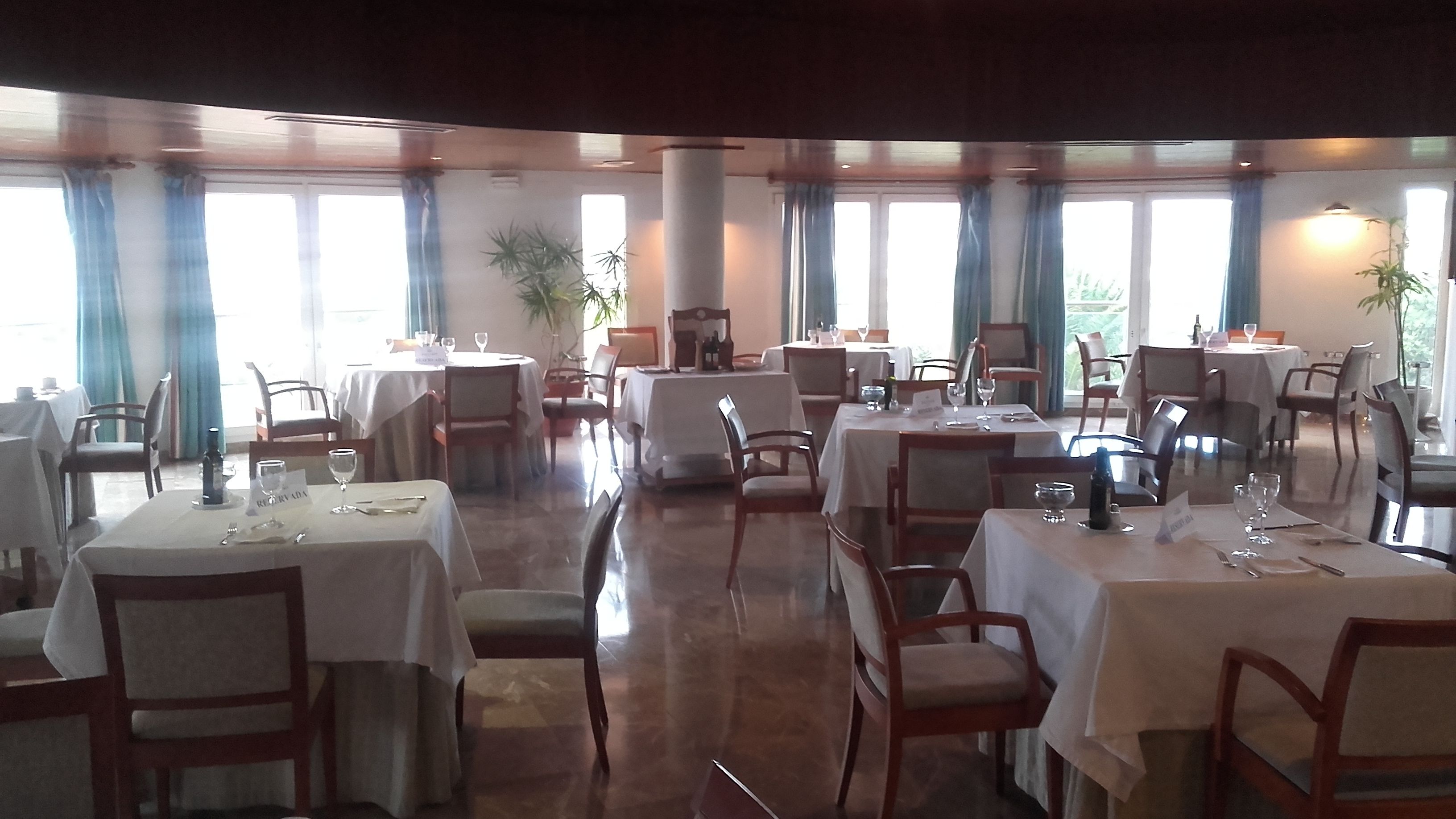 A restaurant with multiple tables and chairs, large windows with curtains, and potted plants.