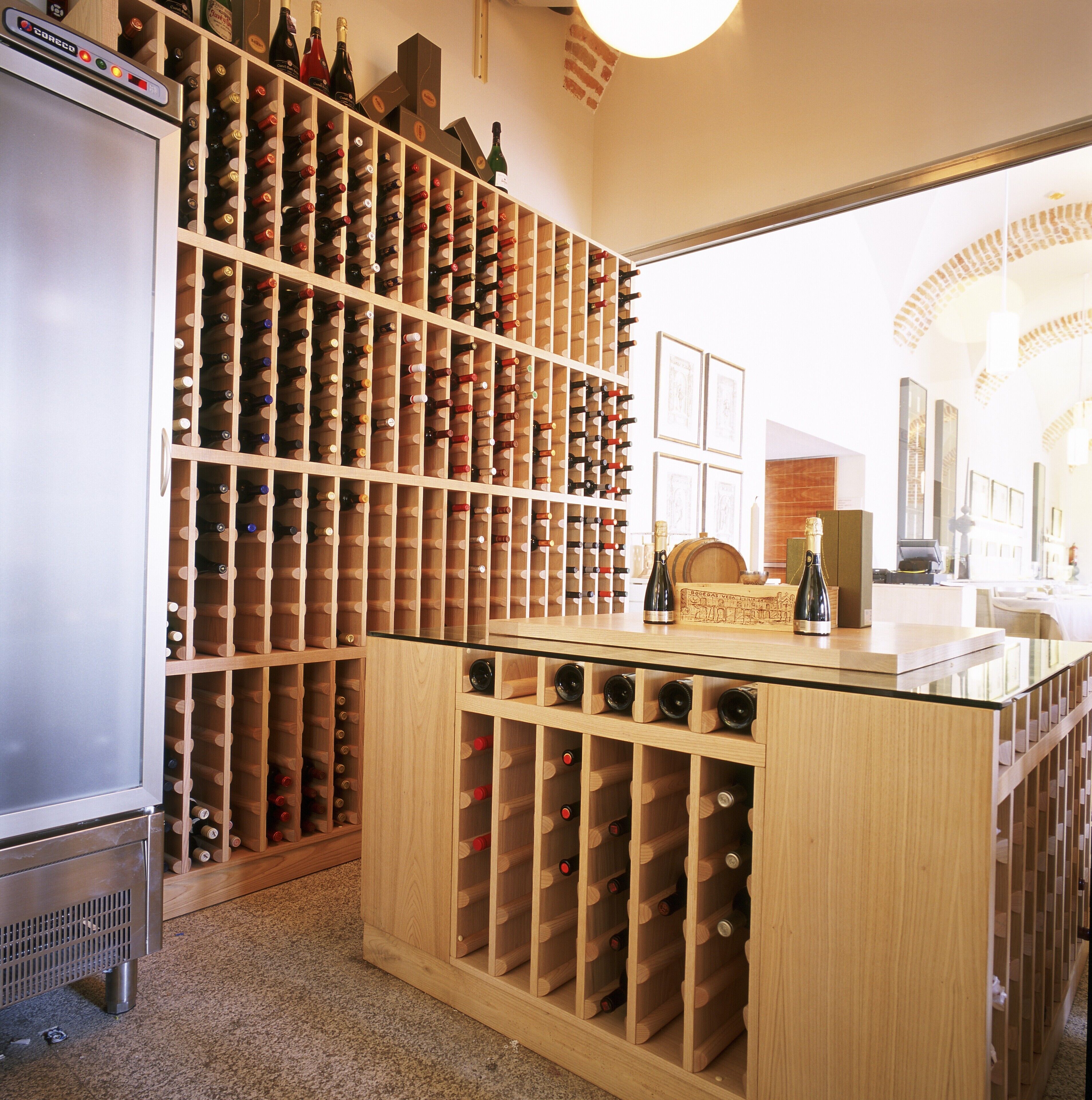 Una bodega con estanterías de madera llenas de botellas de vino, un porta copas de vidrio y un refrigerador.