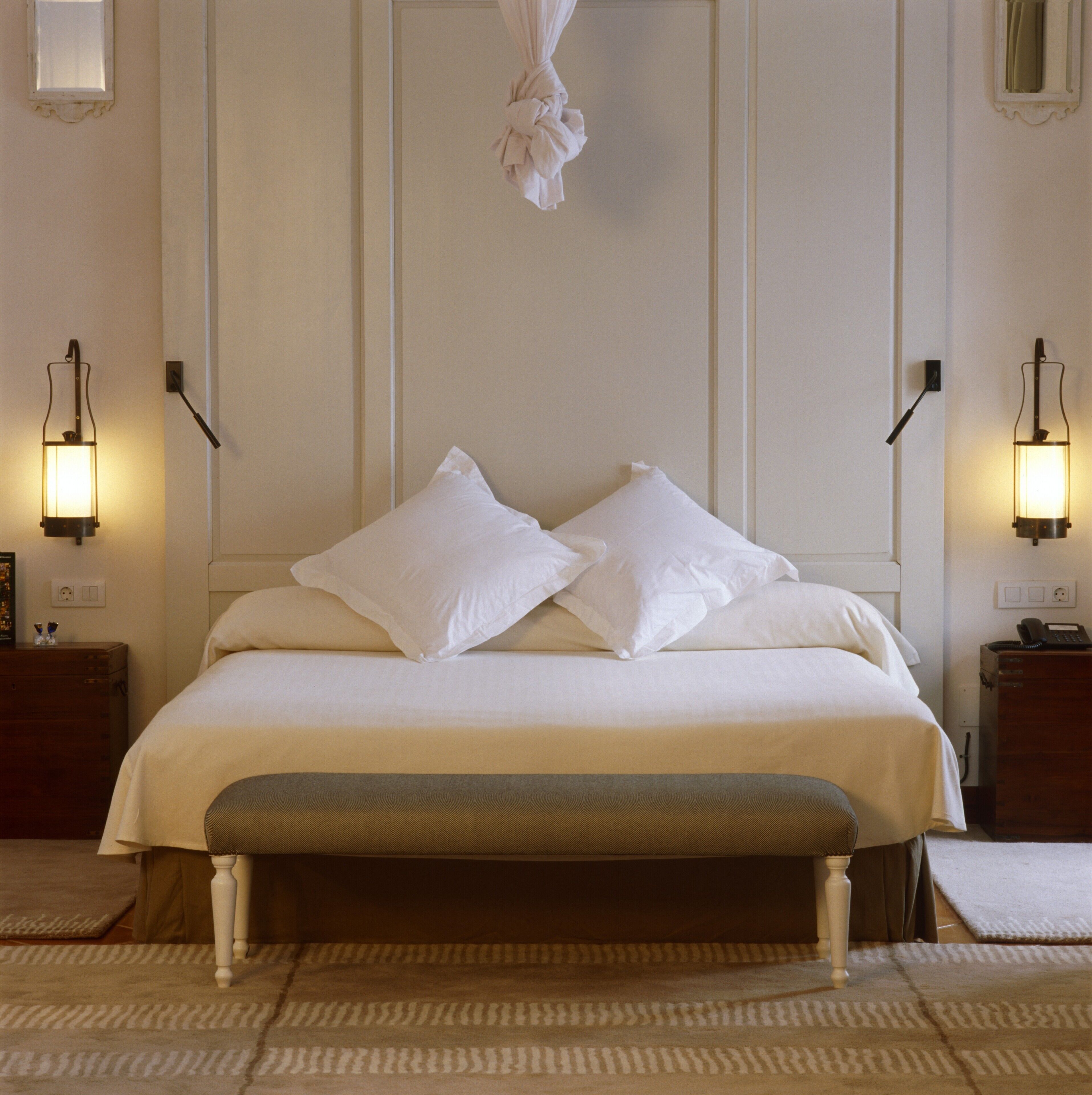 A neatly made bed with white linens and pillows, a bench at the foot of the bed, and two bedside tables with lamps.