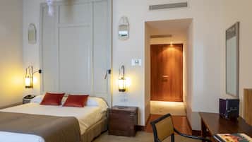 Habitación de hotel con una cama, lámparas de noche, un escritorio con una silla, un armario de madera y una puerta que da a otra habitación.