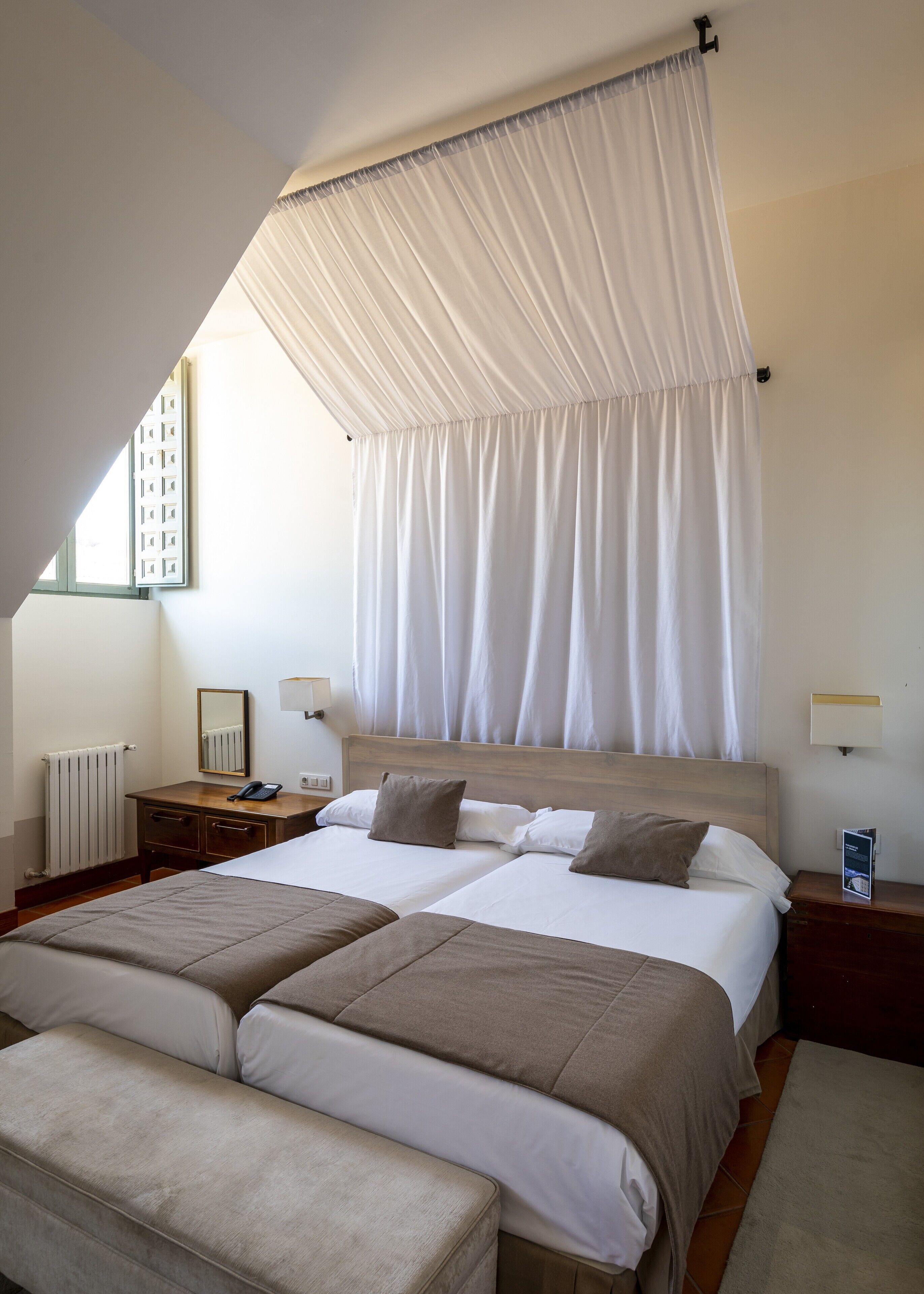 Une chambre d’hôtel avec un grand lit, deux tables de chevet, une petite table, une fenêtre avec des rideaux et un radiateur.