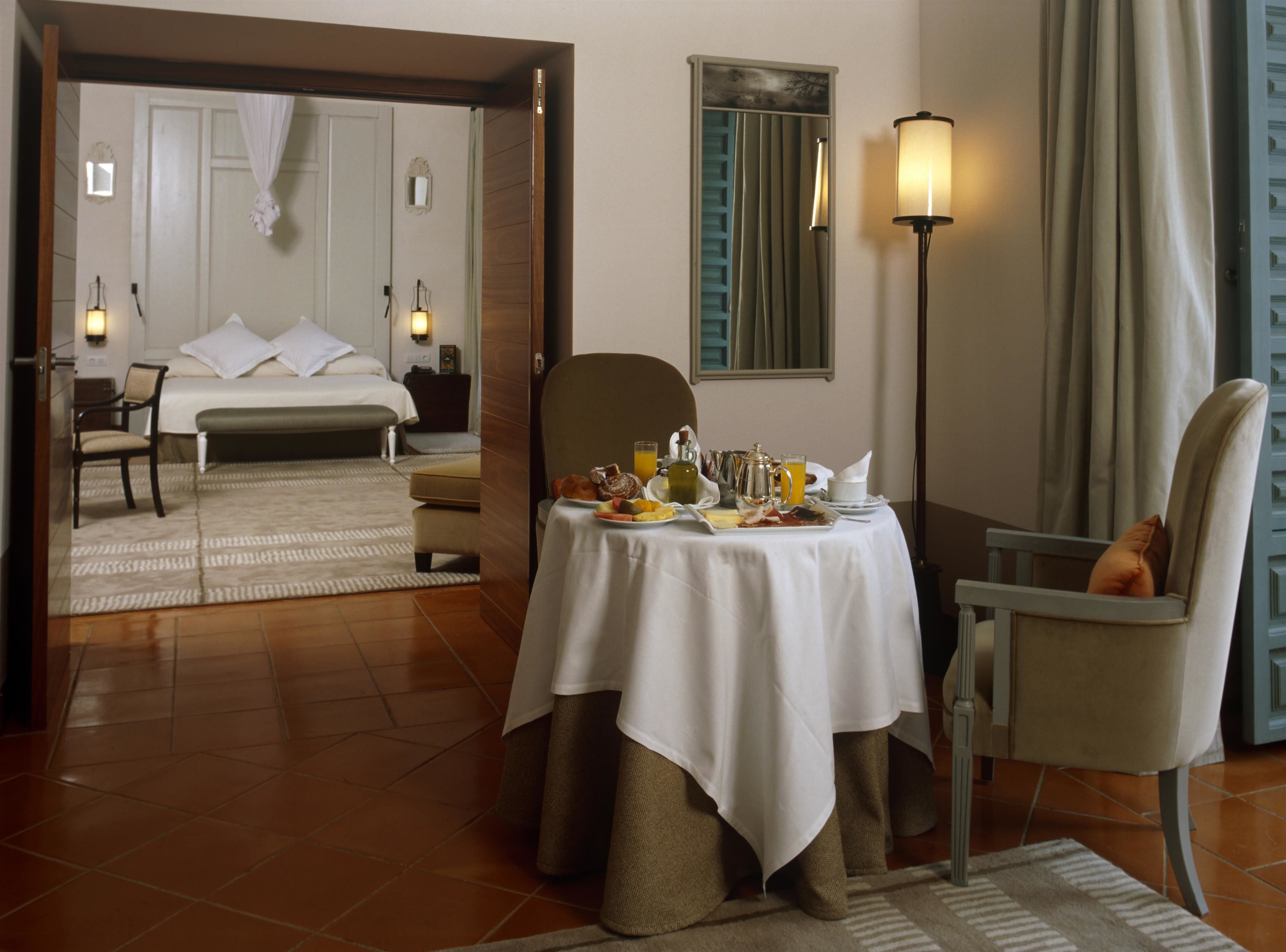 A hotel room with a bed, a dining table with breakfast, a chair, and a lamp.