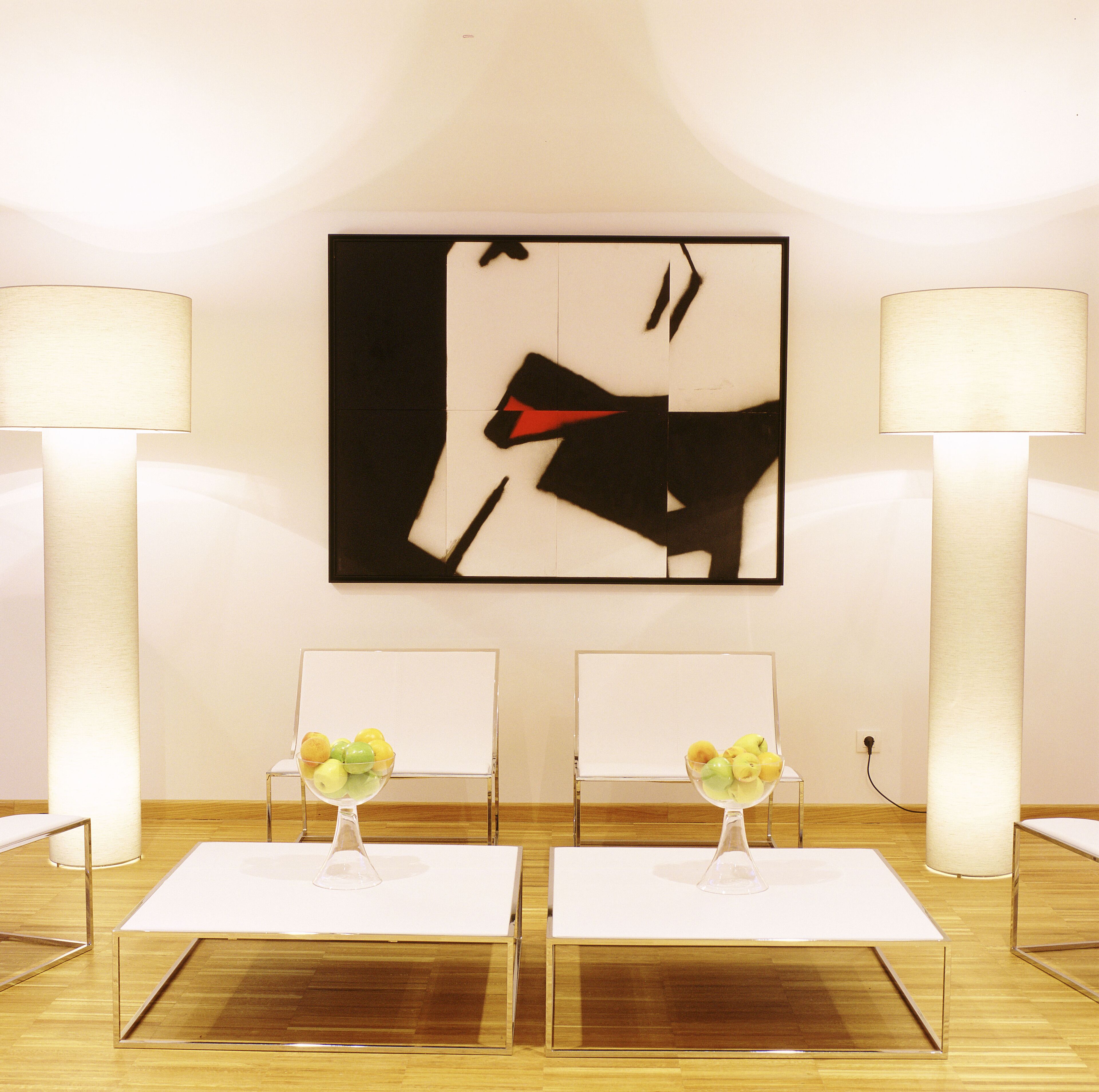 Una habitación moderna con dos sillas blancas, una mesa con un florero de manzanas amarillas y un cuadro abstracto grande en la pared.