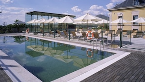 A swimming pool with lounge chairs and umbrellas, surrounded by a wooden deck and glass buildings. - Parador De La Granja (La Granja de San Ildefonso)